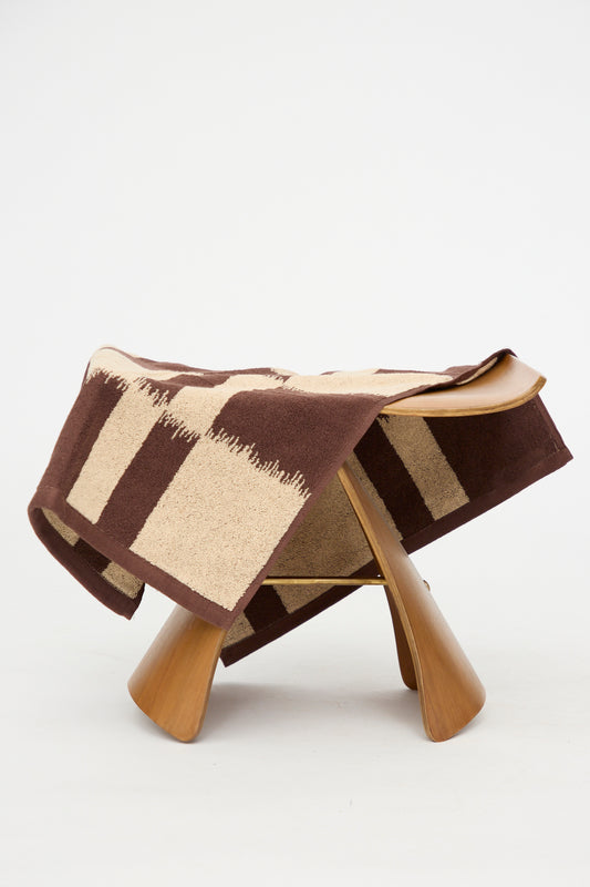 The Karin Hand Towel by Autumn Sonata, featuring a brown and beige checkered Ikat-inspired pattern, is draped over a modern wooden stool with curved legs against a plain white background.