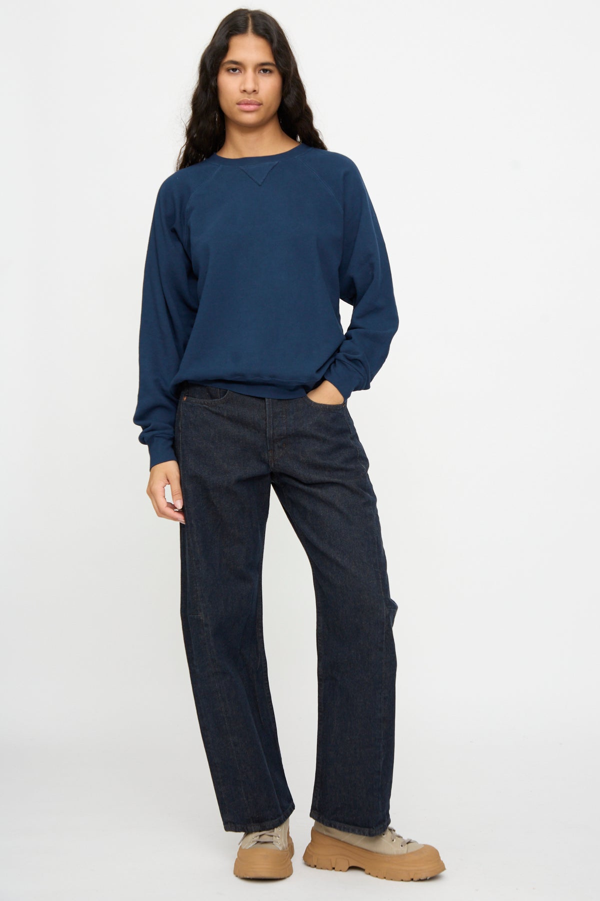 Person stands against a plain background wearing the B Sides Arena Crewneck in Marine Overdye, paired with dark jeans and tan shoes, one hand in their pocket.