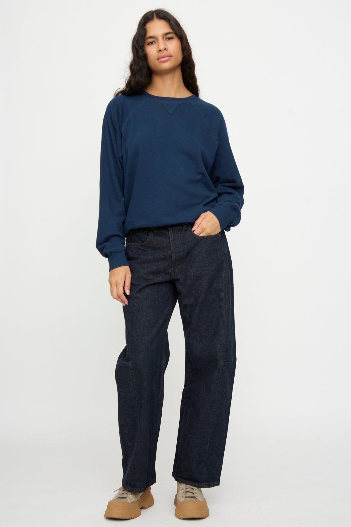 Person stands against a plain background wearing the B Sides Arena Crewneck in Marine Overdye, dark wide-leg jeans, tan shoes, one hand in pocket.