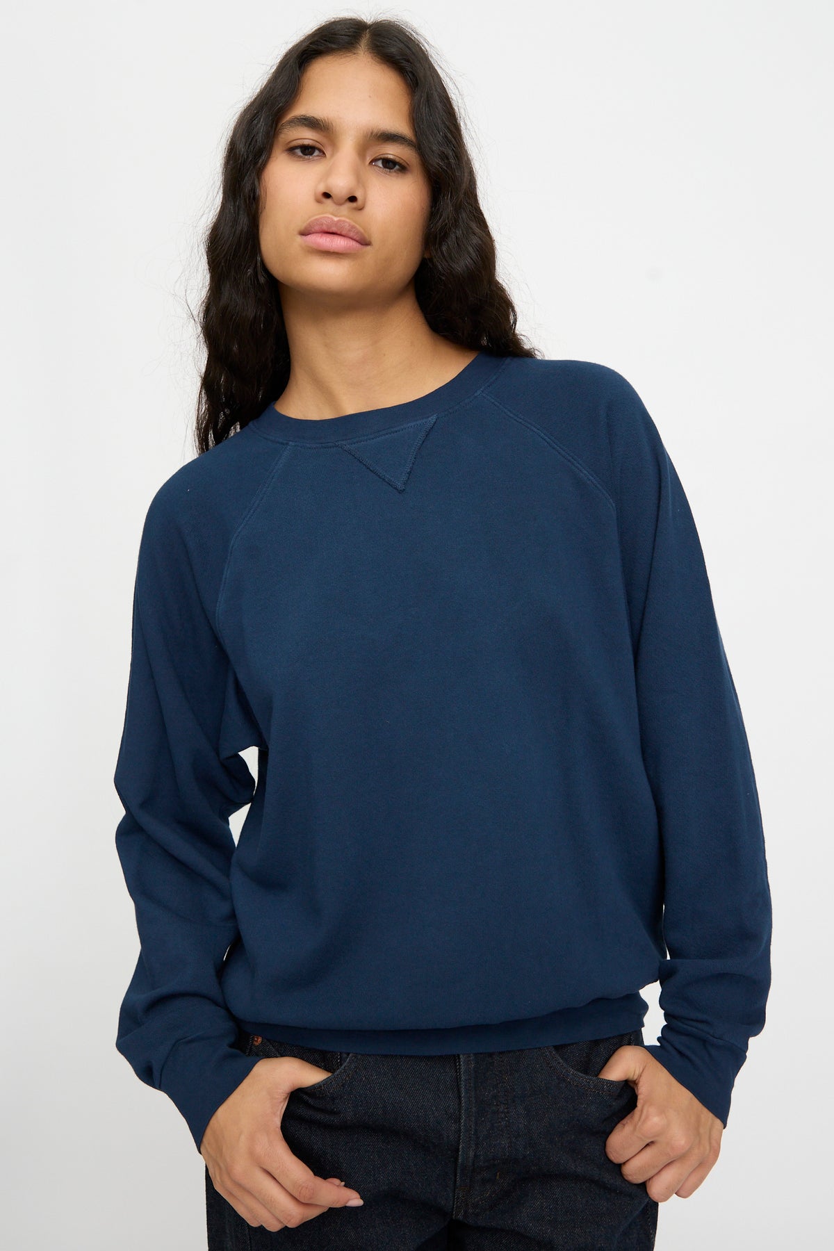 Person with long dark hair wears the B Sides Arena Crewneck in Marine Overdye and dark pants, standing against a plain light background.