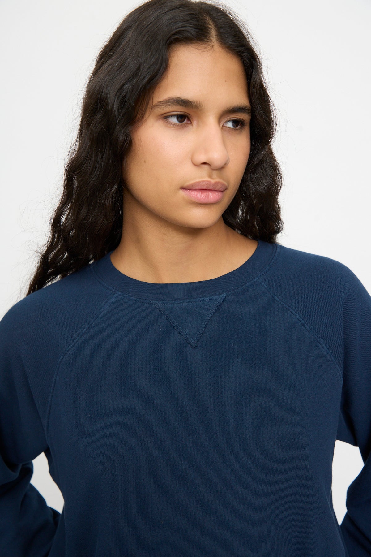 Person with long dark hair wears the B Sides Arena Crewneck in Marine Overdye, relaxed fit, gazing sideways with a neutral expression against a plain background.