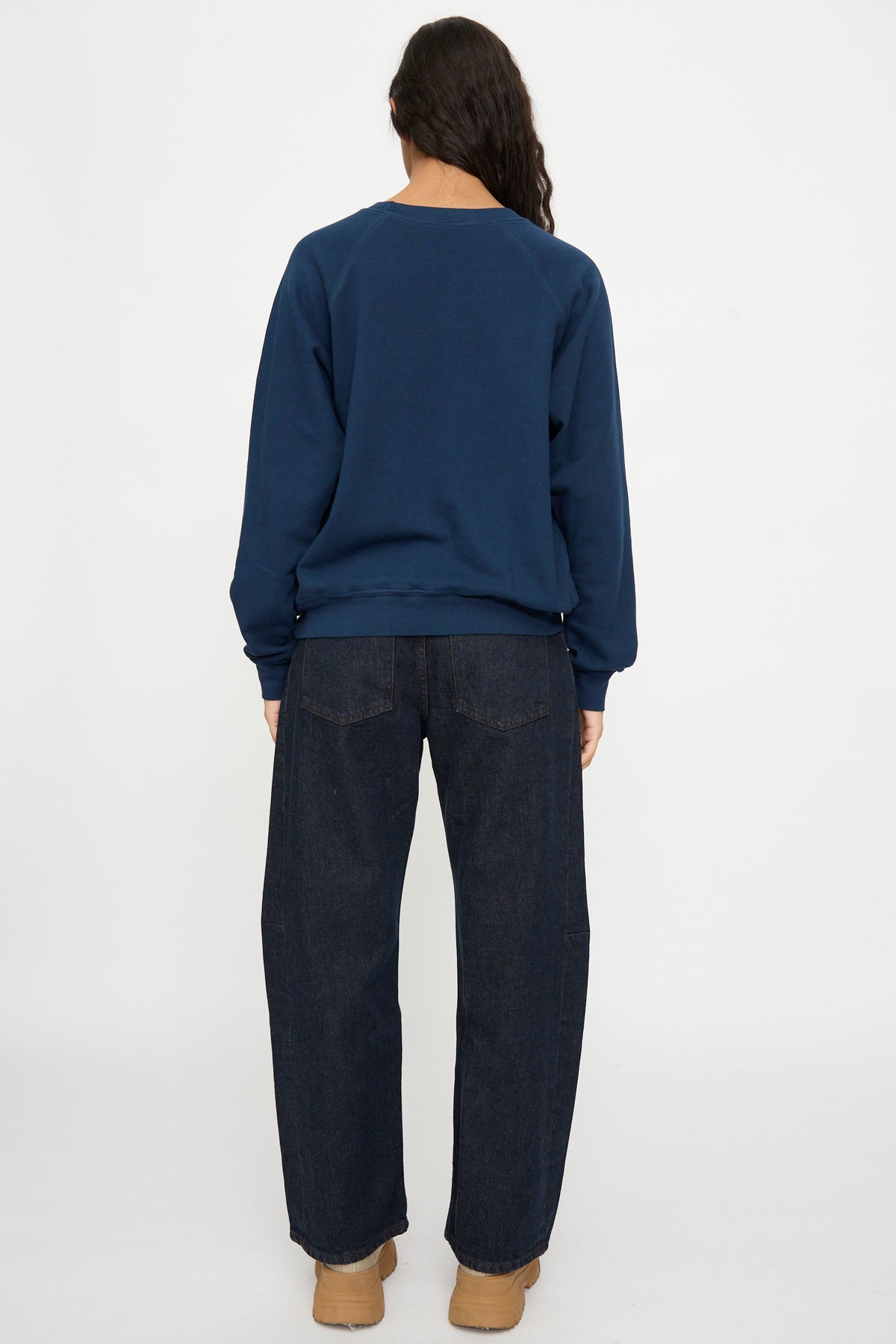 Person faces away, wearing the B Sides Arena Crewneck in Marine Overdye, dark blue jeans, and tan shoes against a plain white background.