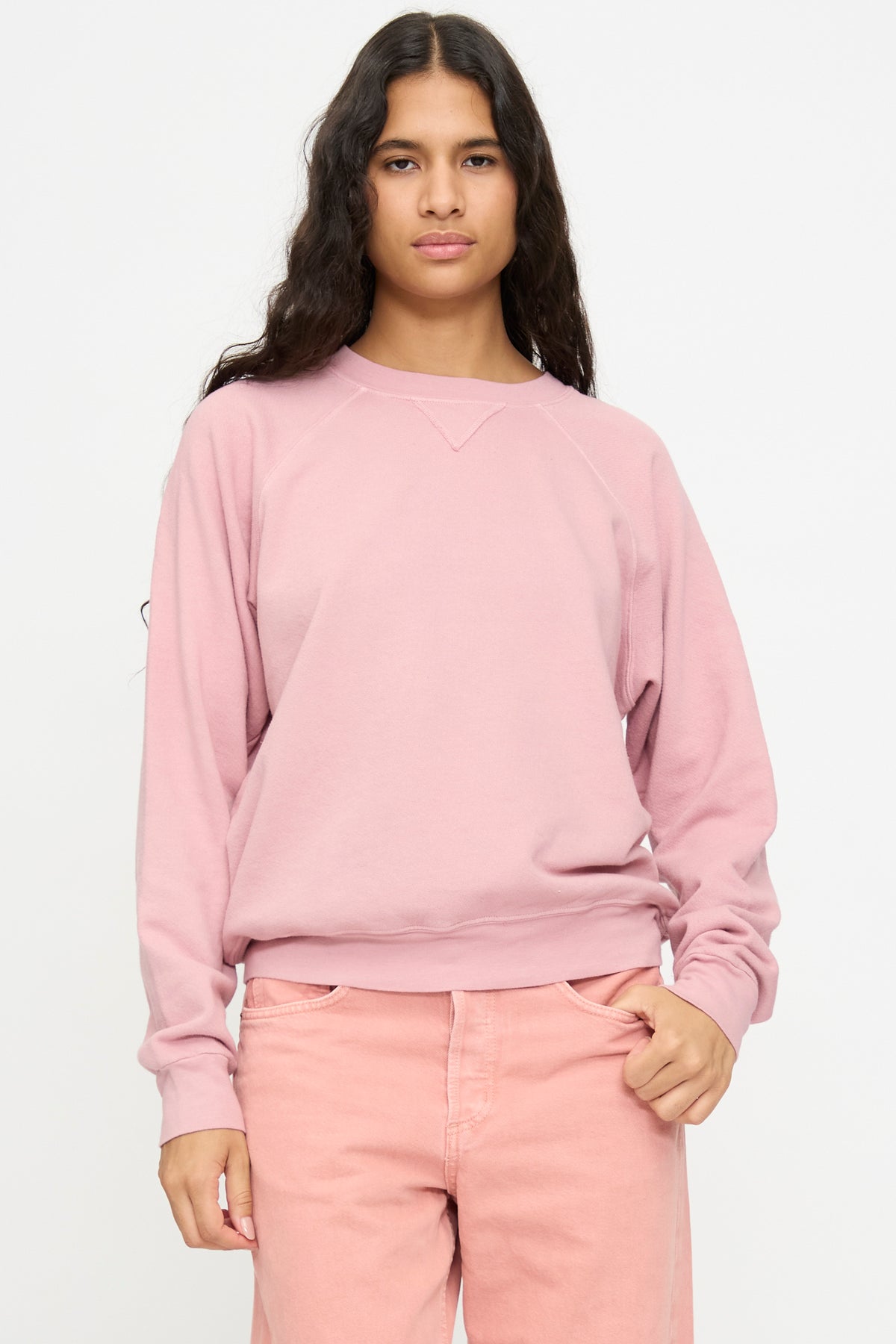 Person with long dark hair wears the Arena Crewneck in Pink Overdye by B Sides, paired with matching pants, standing against a plain white background.