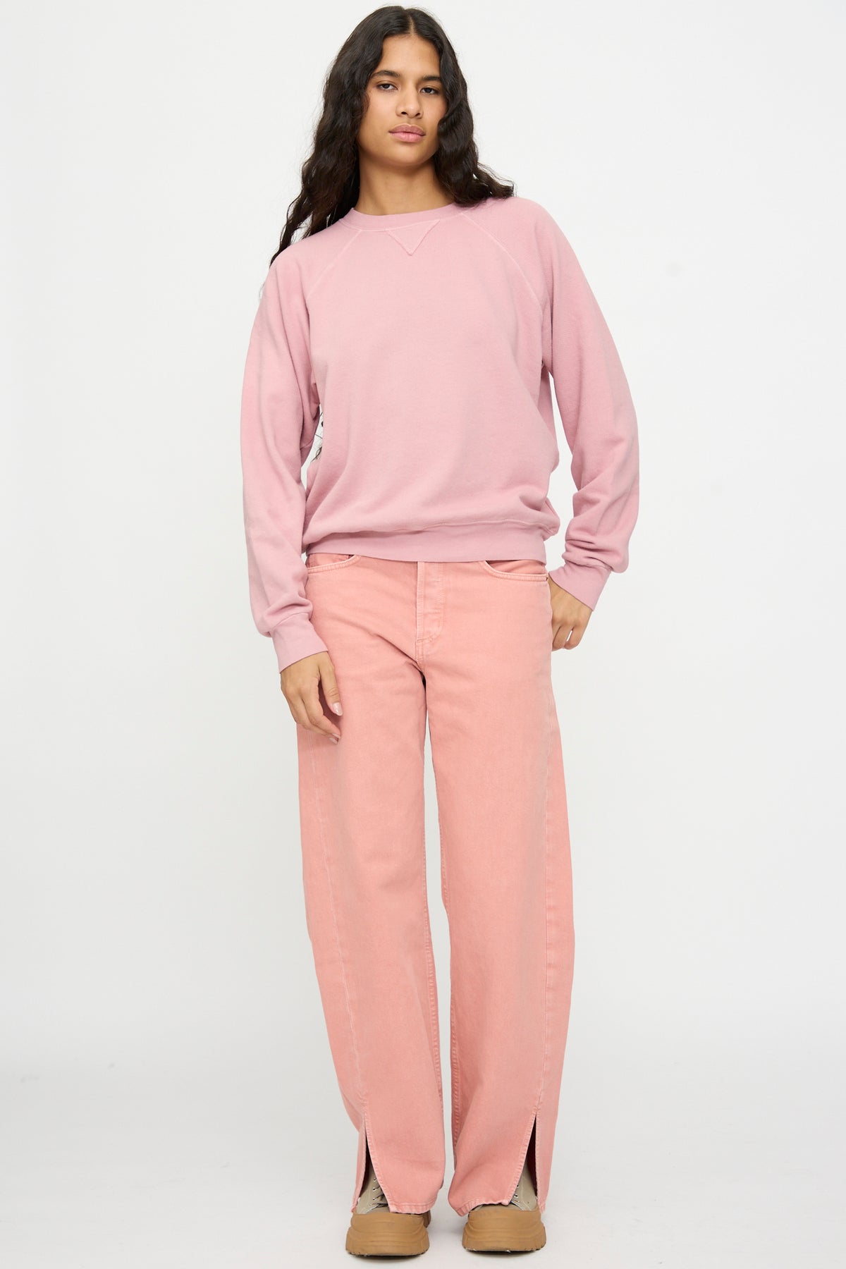 Someone stands against a plain background wearing the B Sides Arena Crewneck in Pink Overdye, pink wide-leg jeans with front slits, and tan shoes.
