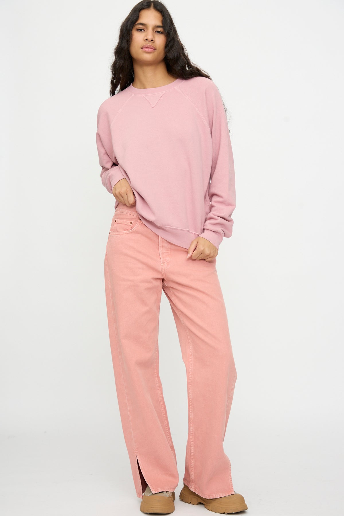 Person stands against a plain background, wearing the Arena Crewneck in Pink Overdye by B Sides and matching wide-leg pink pants with slit hems, plus tan shoes.