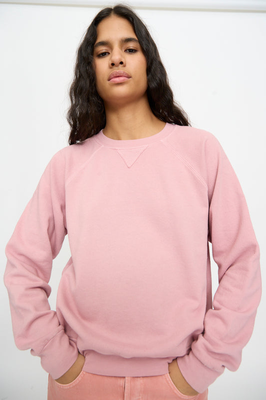 Person with long dark hair wears the B Sides Arena Crewneck in Pink Overdye and matching pants, standing against a plain white background with hands in pockets.
