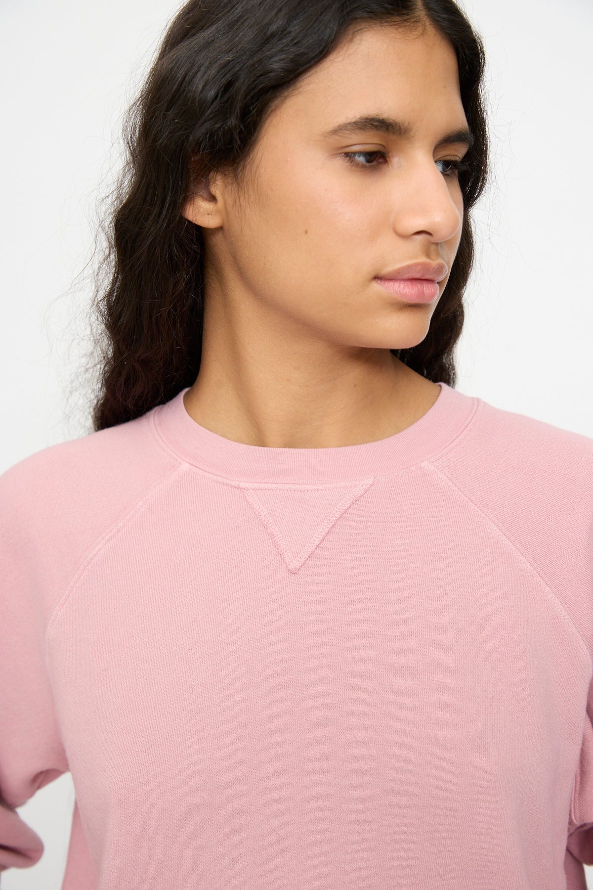 Person with long, wavy dark hair wears the B Sides Arena Crewneck in Pink Overdye, looking to the side against a plain background.