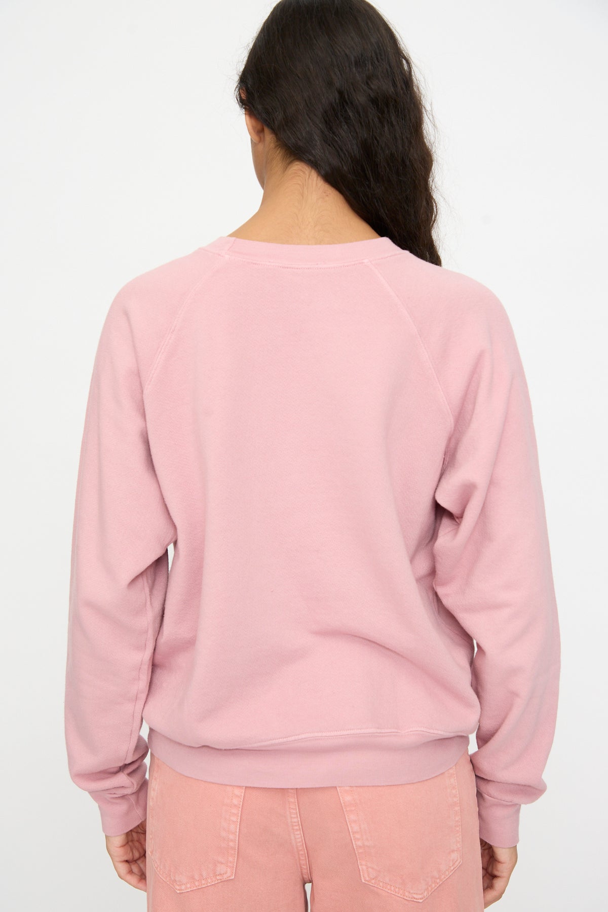 Person with long dark hair wears the Arena Crewneck in Pink Overdye by B Sides and peach-colored jeans, shown from the back against a plain white background.