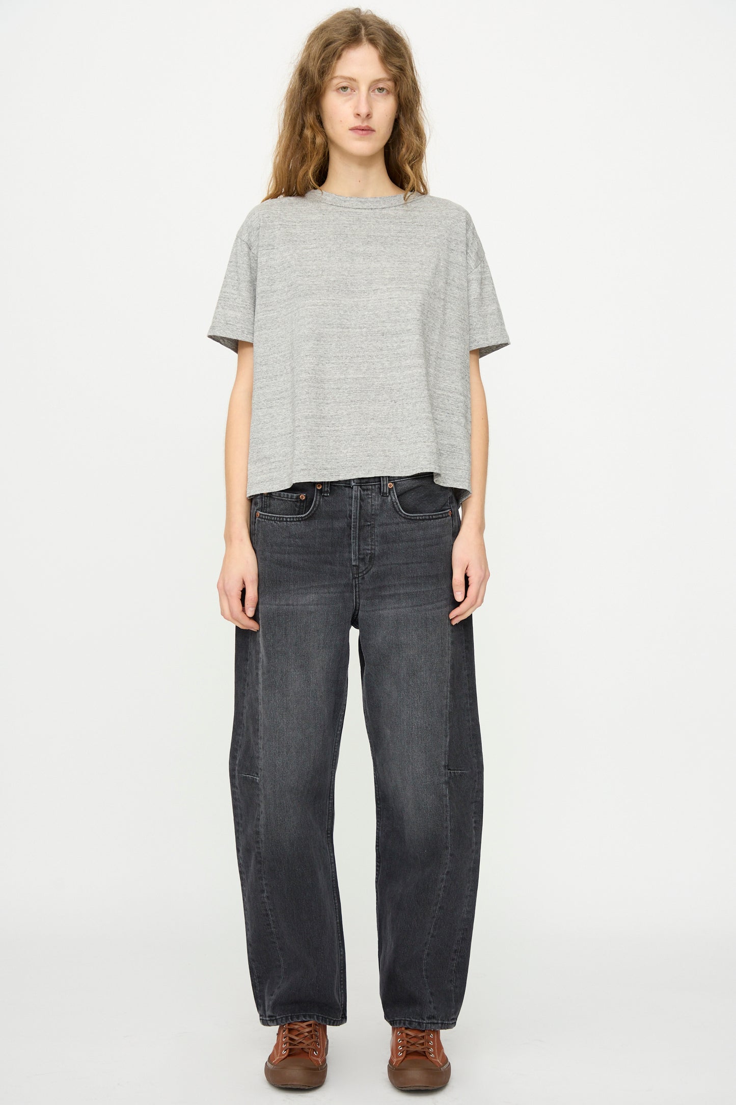 A person stands facing forward wearing the B Sides Atlas Tee in Heather Grey, paired with dark baggy jeans and brown lace-up shoes against a plain white background.