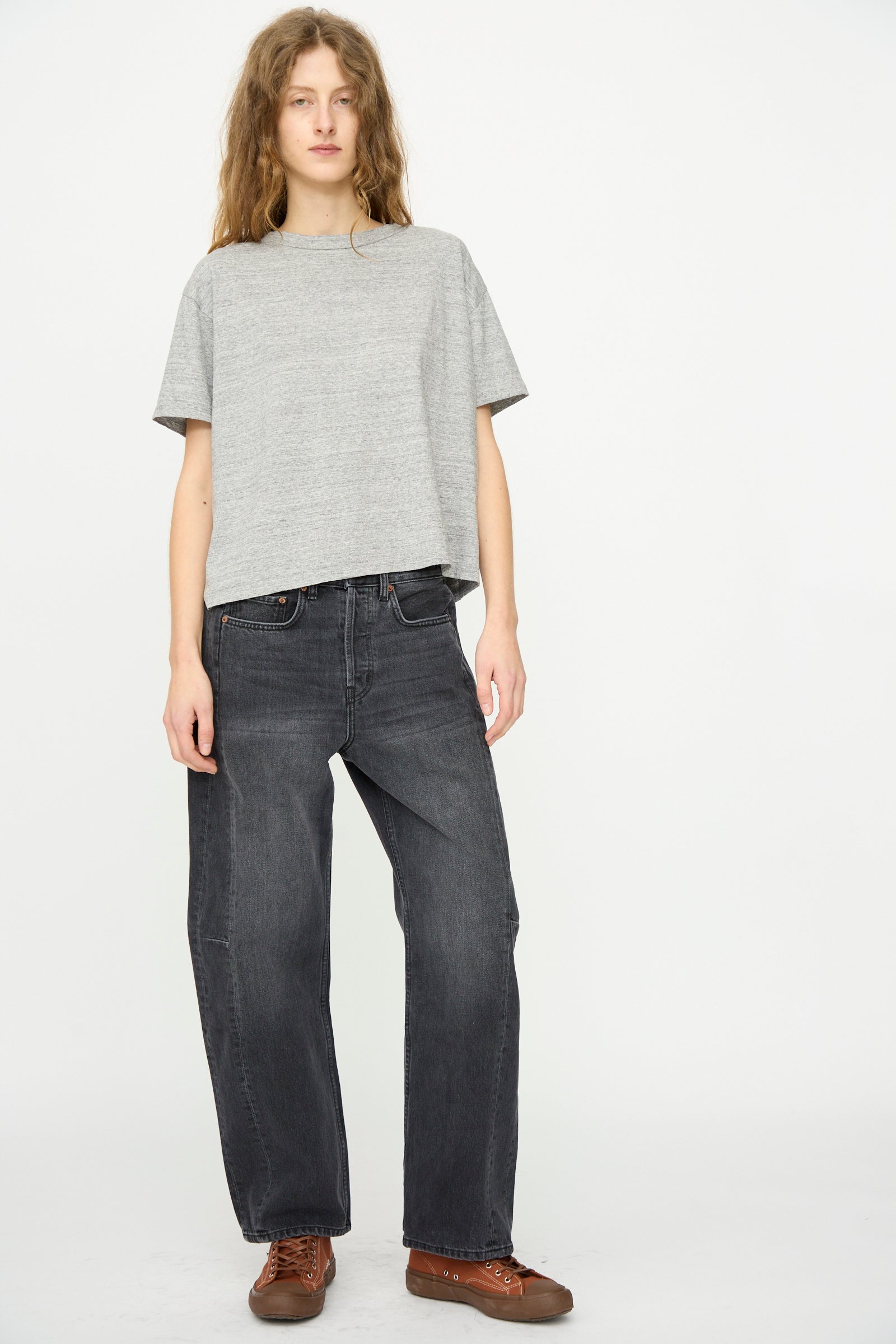 A person stands against a plain white background, wearing the B Sides Atlas Tee in Heather Grey with dark baggy jeans and brown shoes.