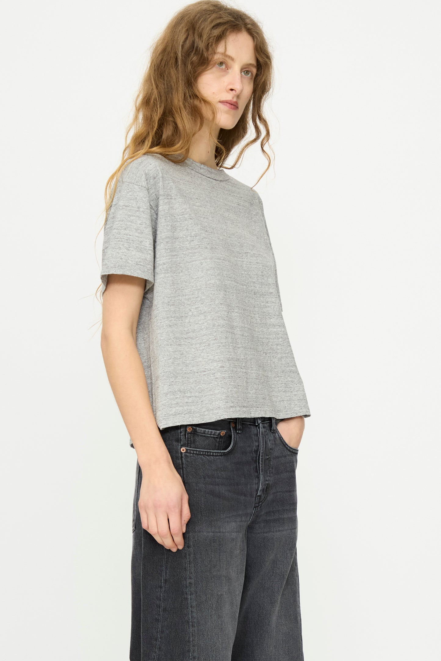 A person with long wavy hair wears the B Sides Atlas Tee in Heather Grey and dark jeans, standing against a plain white background.
