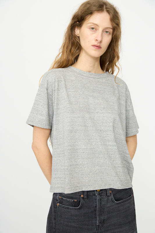 A person with long wavy hair wears the B Sides Atlas Tee in Heather Grey and black jeans, standing against a plain white background.