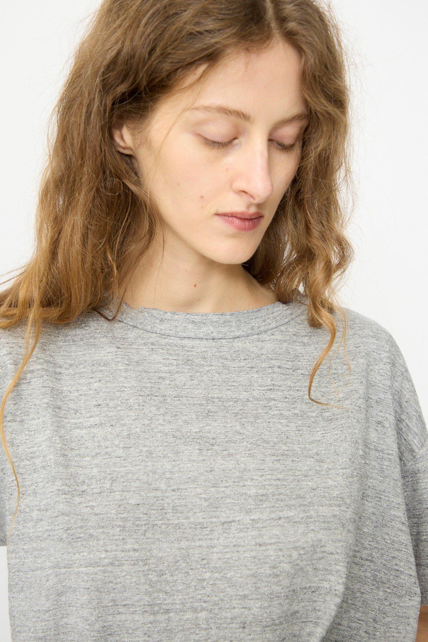 A person with long wavy hair wears the B Sides Atlas Tee in Heather Grey, a relaxed fit made of Japanese milled cotton, looking downward with a neutral expression.