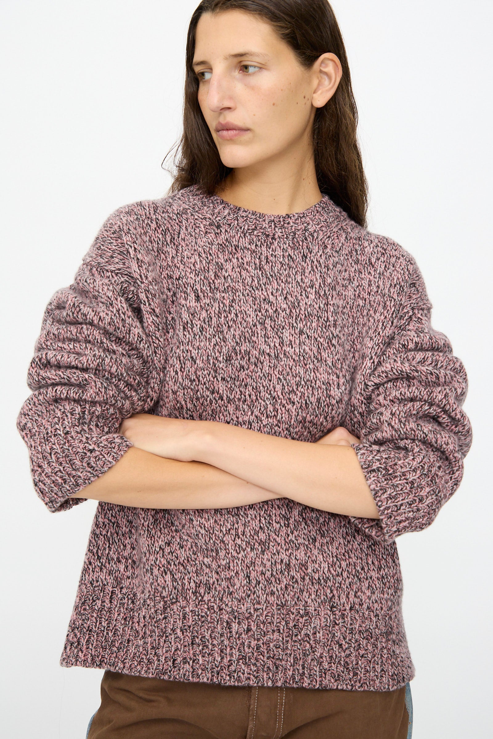 A person with long brown hair wears the B Sides Braided Lambswool Crew in Etowah Twist, a marled pink and black lambswool sweater, paired with brown pants, standing with arms crossed against a plain background.