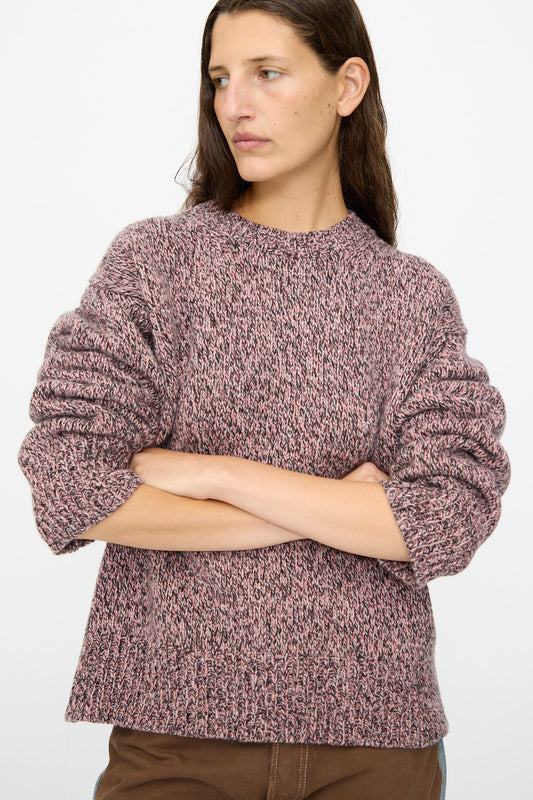A person with long brown hair wears the B Sides Braided Lambswool Crew in Etowah Twist, a marled pink and black lambswool sweater, paired with brown pants, standing with arms crossed against a plain background.