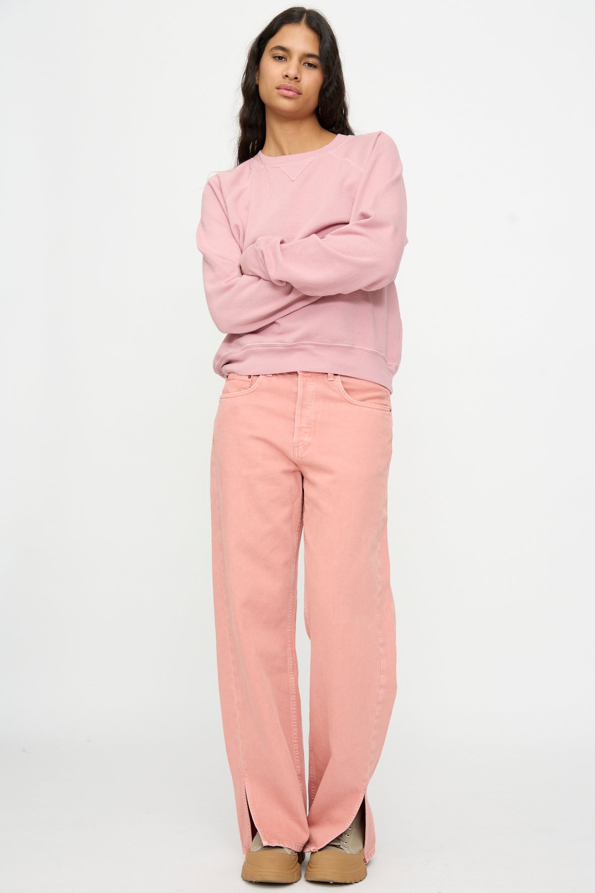 Person standing with arms crossed, wearing a light pink sweatshirt and B Sides Charlot Split Hem in Myra Pink Overdye jeans, paired with tan shoes against a plain white background.