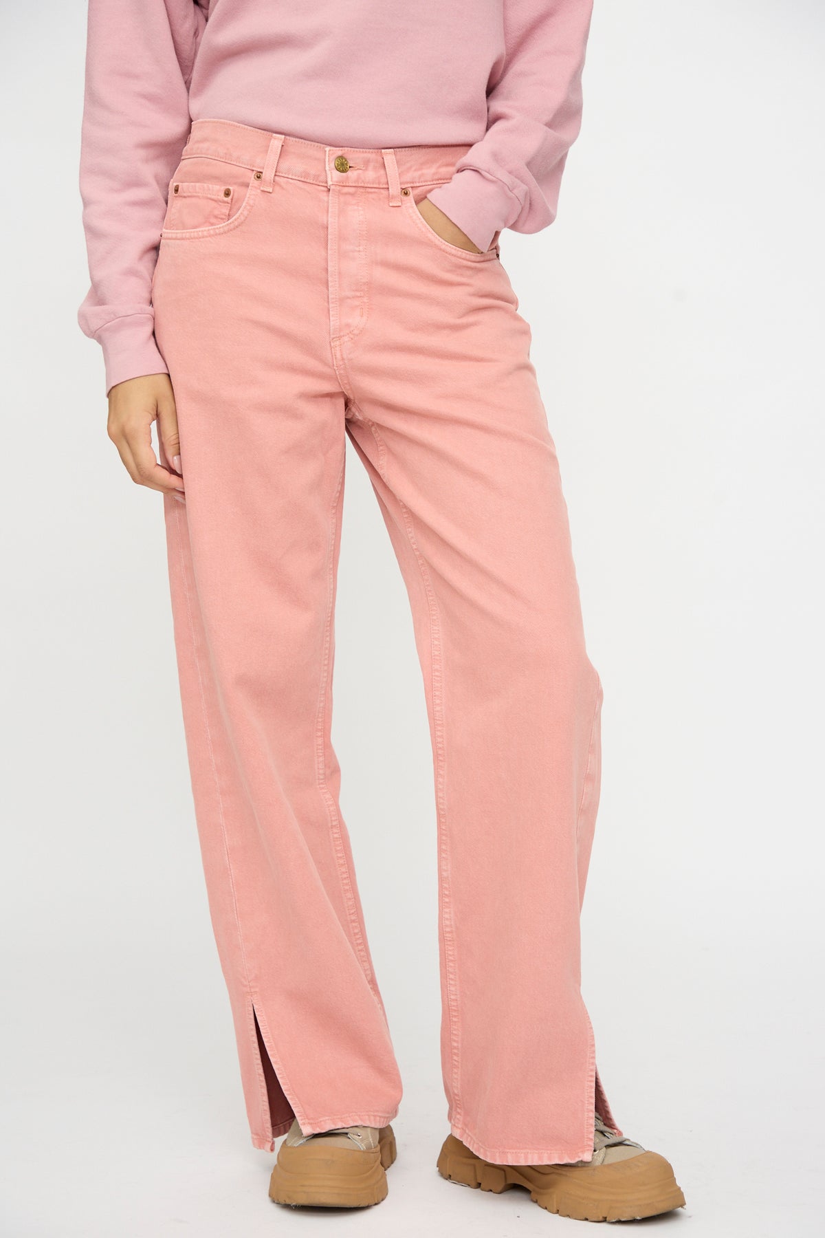Person wears Charlot Split Hem in Myra Pink Overdye by B Sides, a pink sweatshirt, and tan shoes. Left hand in pocket. Plain white background.