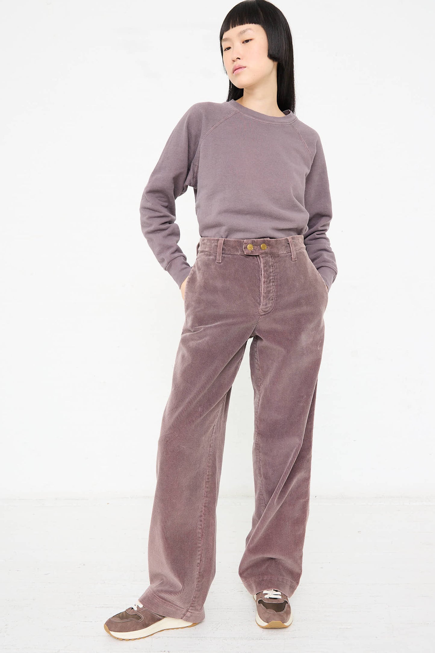 A person dressed in a mauve ensemble stands against a plain white background, wearing a long-sleeve top and B Sides' Corduroy Chino in Tess Overdye paired with sneakers.