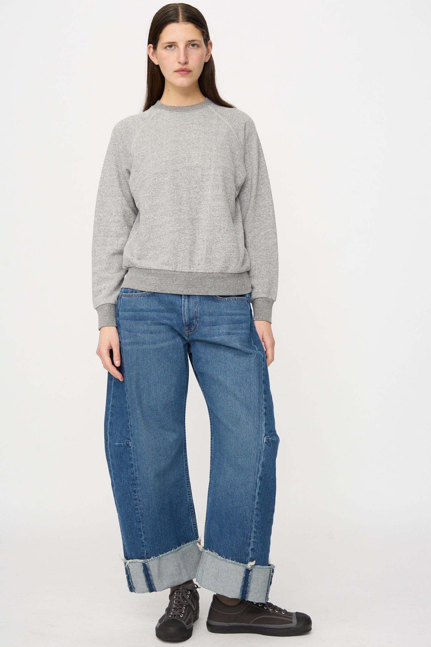 A person stands facing forward against a white background, wearing the B Sides Crewneck Sweater in Heather Grey with wide-leg cuffed blue jeans and dark sneakers.