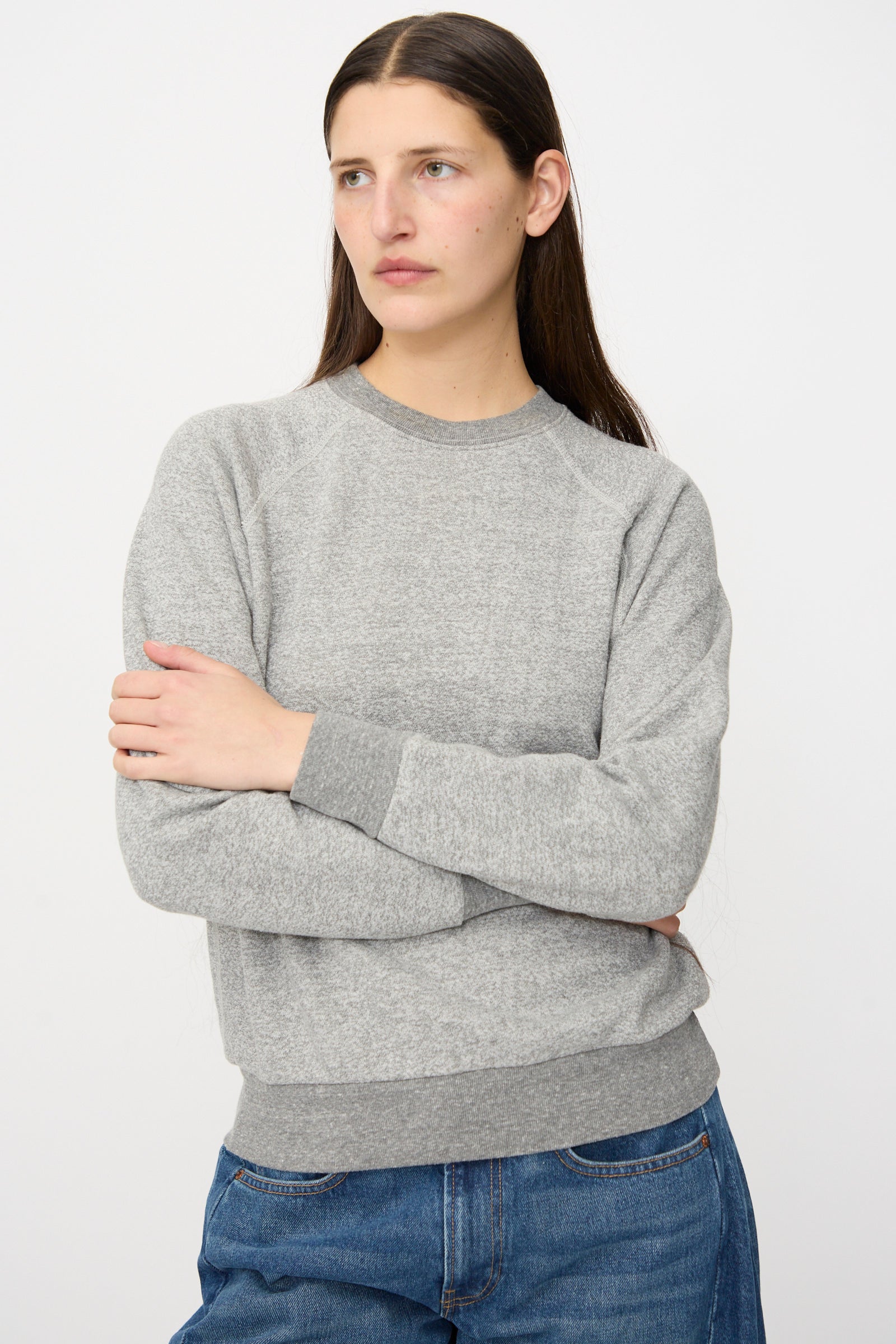 A person with long brown hair wears the B Sides Crewneck Sweater in Heather Grey and blue jeans, standing with arms crossed against a plain white background.