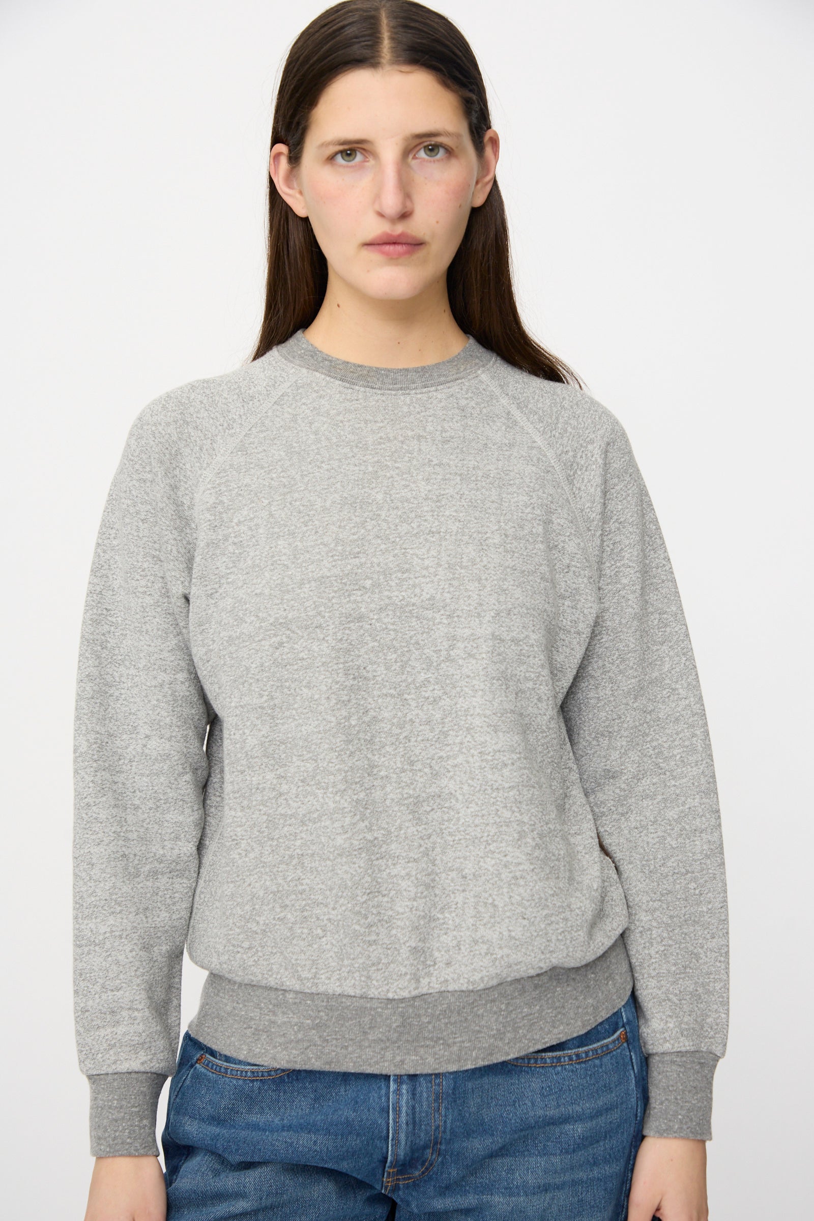 A person with long brown hair wears the B Sides Crewneck Sweater in Heather Grey and blue jeans, standing against a plain white background.
