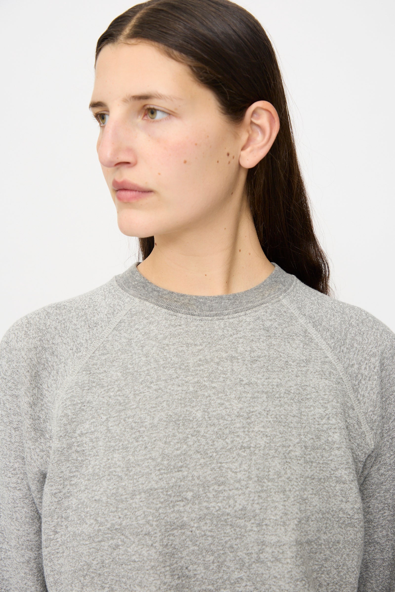 A person with long brown hair models the B Sides Crewneck Sweater in Heather Grey, a relaxed fit style, while looking to the side against a plain light background.
