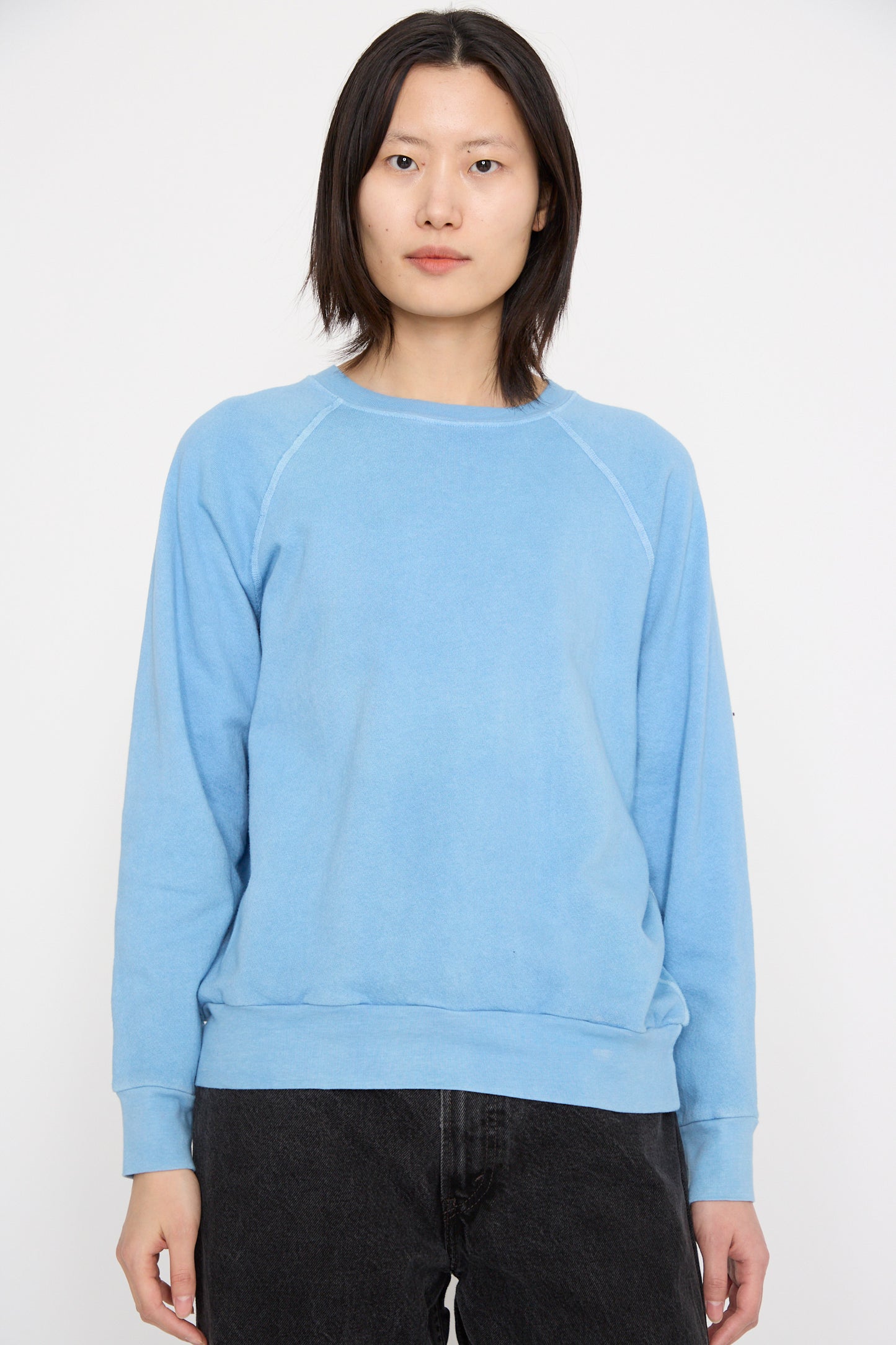 A model wearing B Sides' Crewneck Sweatshirt in Arlo Blue, featuring raglan sleeves, paired with dark pants stands against a plain white background.