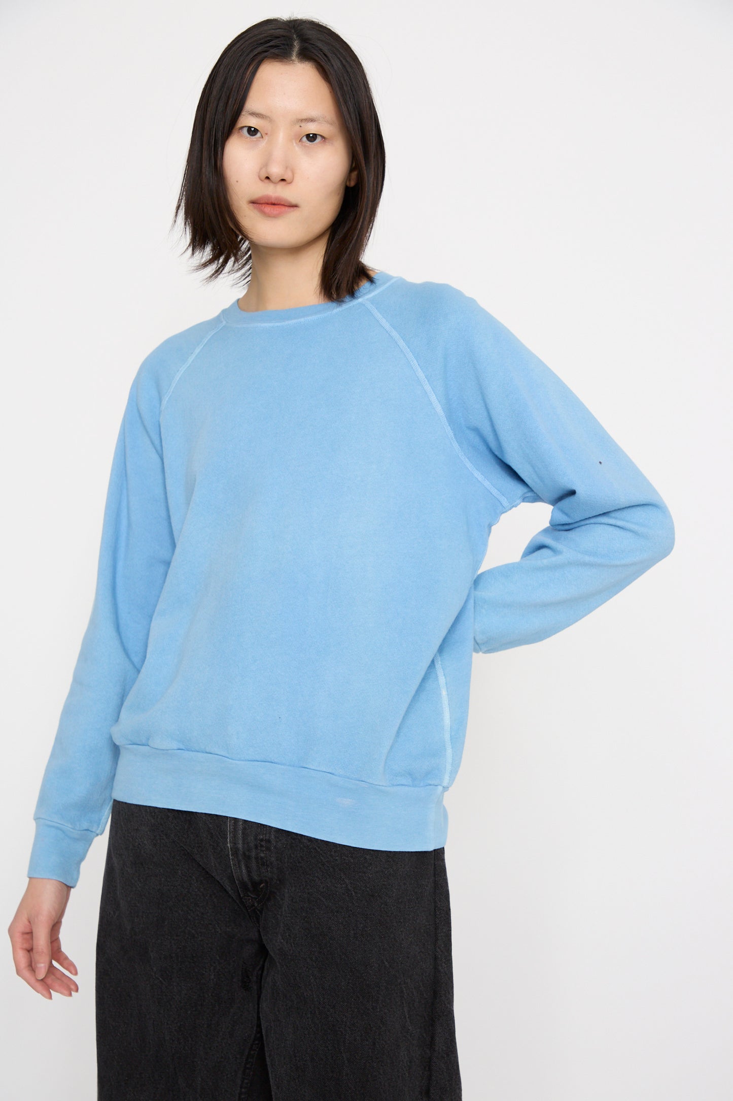 A person is wearing the B Sides Crewneck Sweatshirt in Arlo Blue with raglan sleeves and dark pants, standing against a plain white background with a hand on their hip.