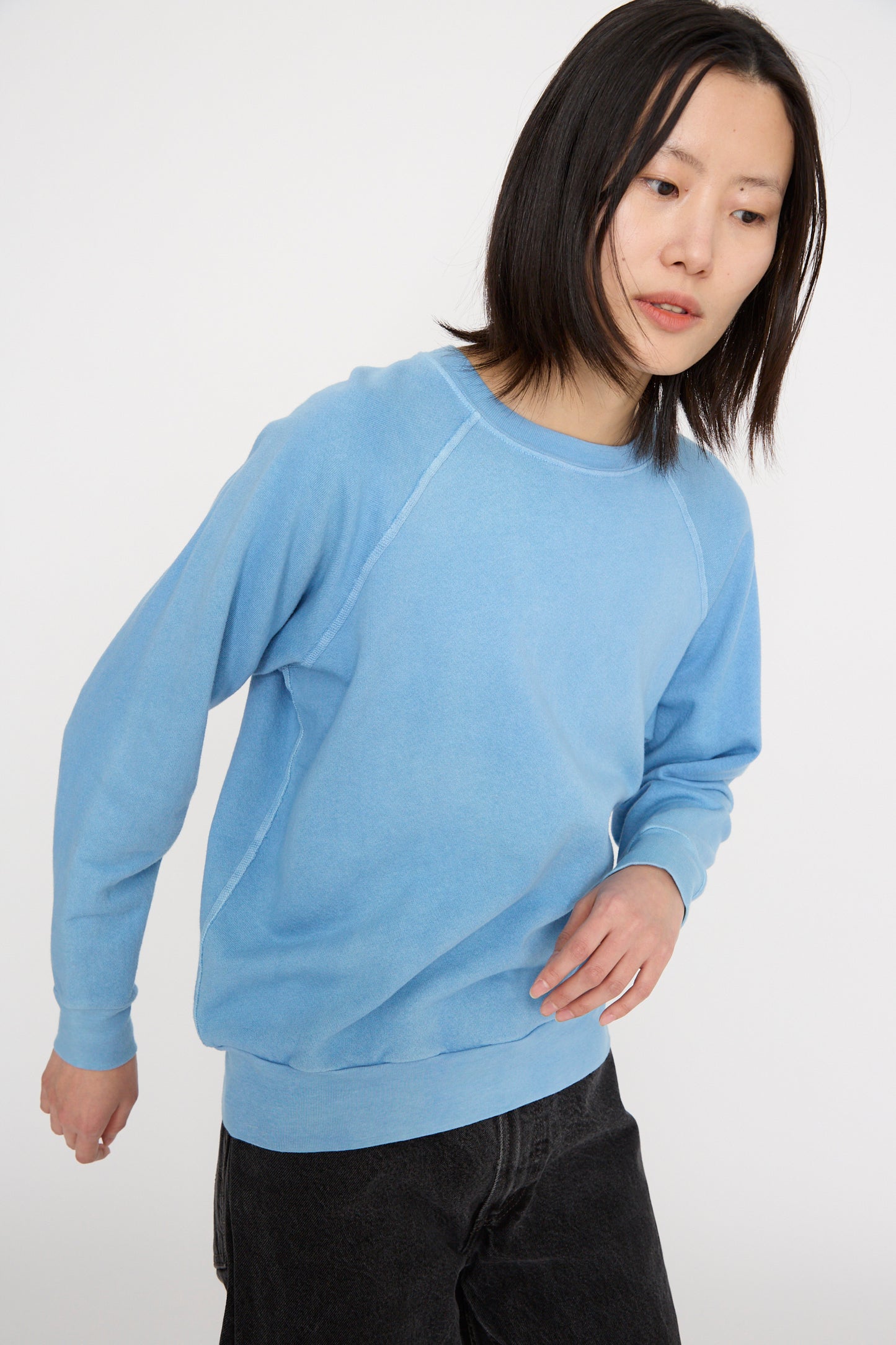 With shoulder-length hair, a person models the B Sides Crewneck Sweatshirt in Arlo Blue, featuring raglan sleeves, paired with dark pants, against a plain white background.