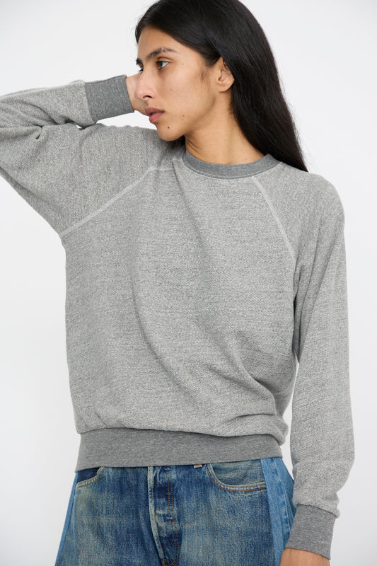 A person with long dark hair wears the B Sides Crewneck Sweatshirt in Heather Grey and blue jeans, looking to the side with one hand behind their head against a neutral background.