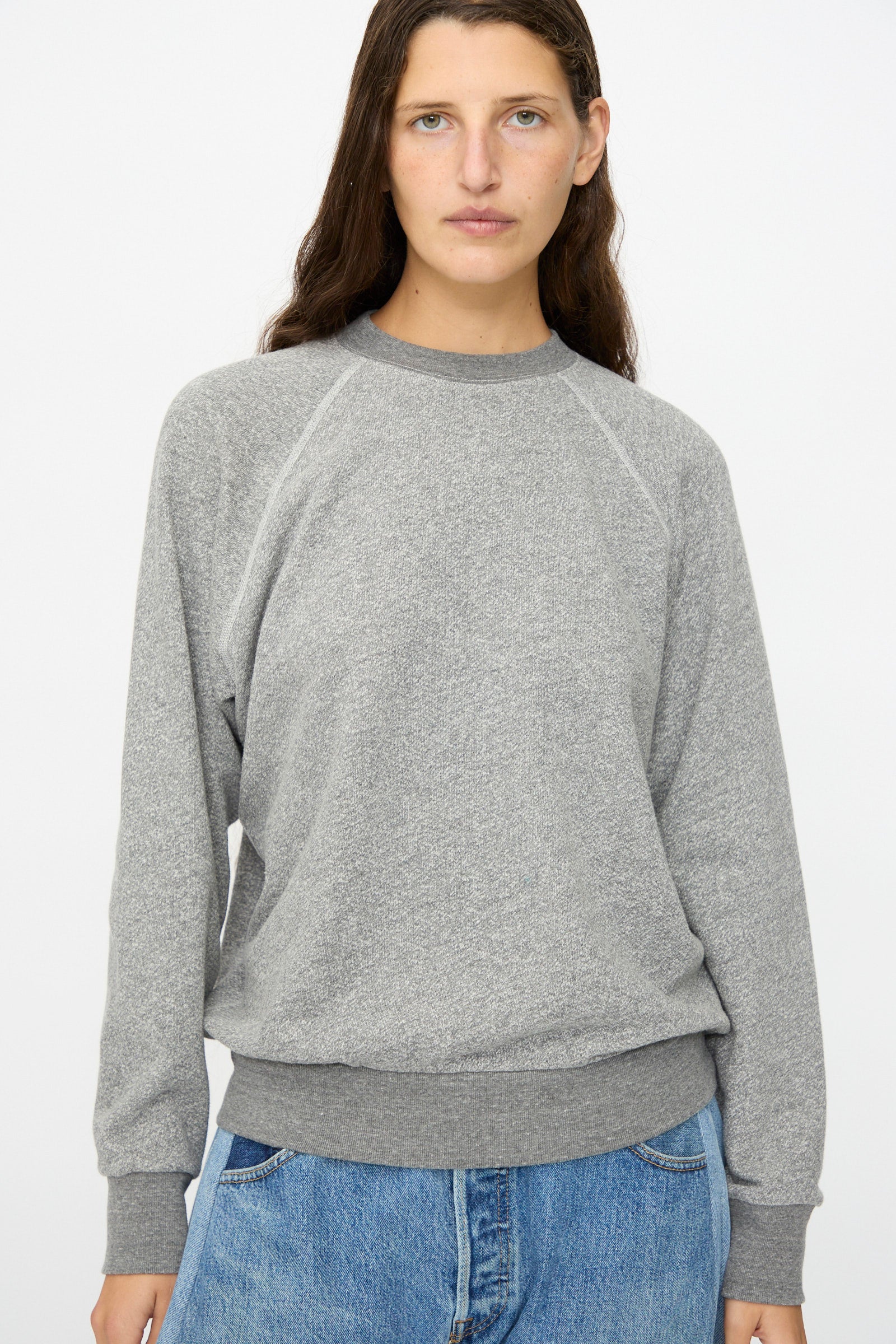 A person with long brown hair wears the B Sides Crewneck Sweatshirt in Heather Grey and blue jeans, standing against a plain white background.