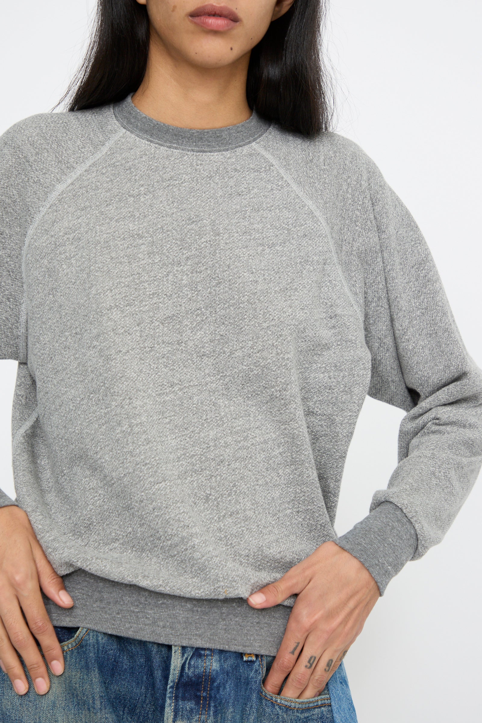 A person wearing a Crewneck Sweatshirt in Heather Grey by B Sides and blue jeans stands against a plain background, with hands reaching for their pockets.