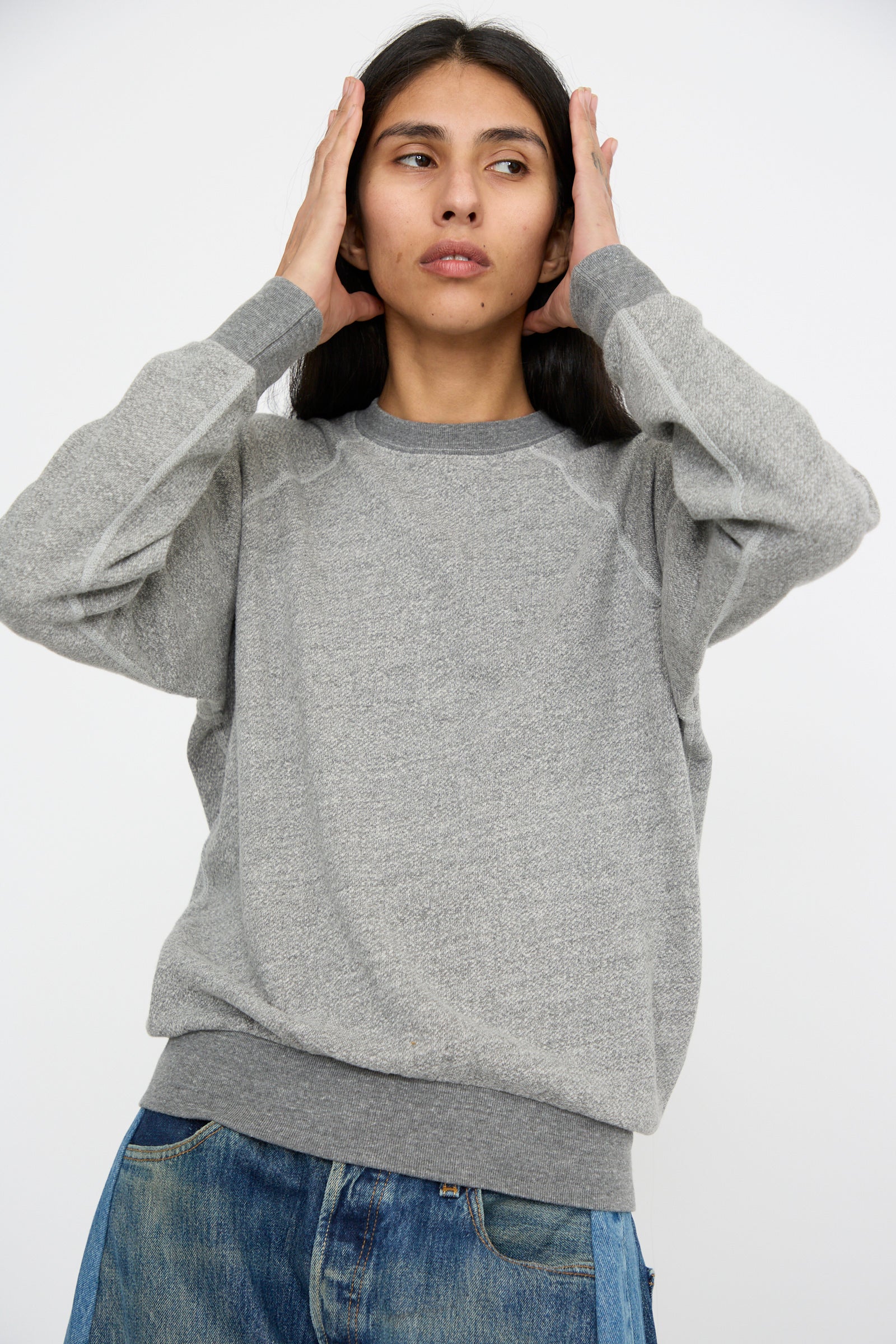 A person in a Crewneck Sweatshirt in Heather Grey by B Sides and jeans stands against a plain background, hands holding their head. 