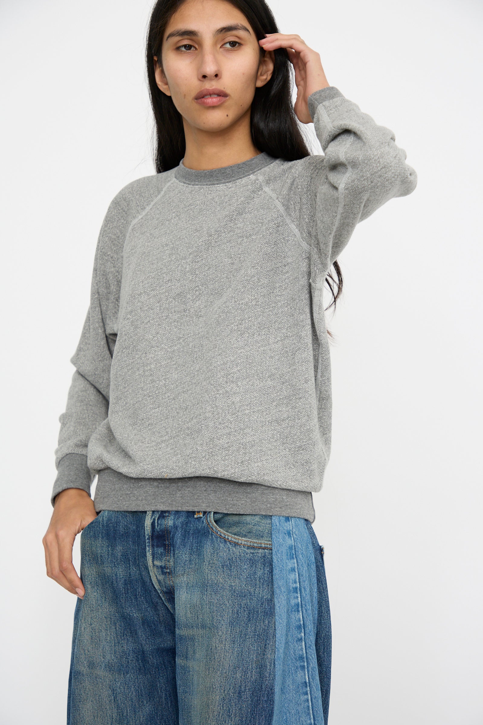 A person dressed in a Crewneck Sweatshirt in Heather Grey by B Sides and blue jeans stands against a plain background with one arm raised.