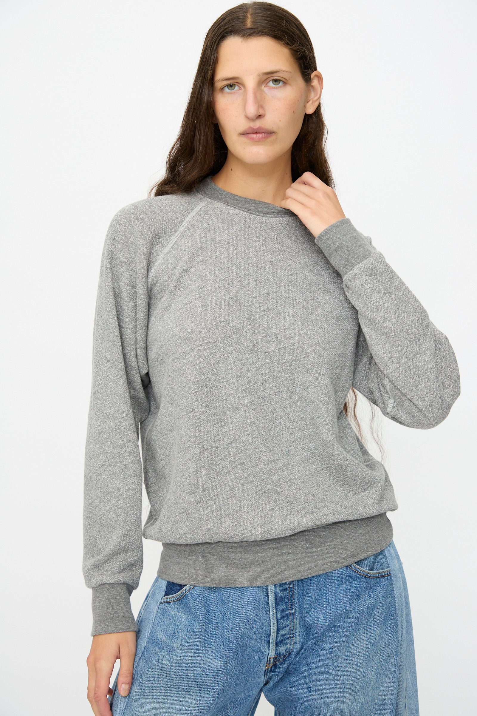 A person with long brown hair wears the B Sides Crewneck Sweatshirt in Heather Grey and blue jeans, standing against a plain white background.