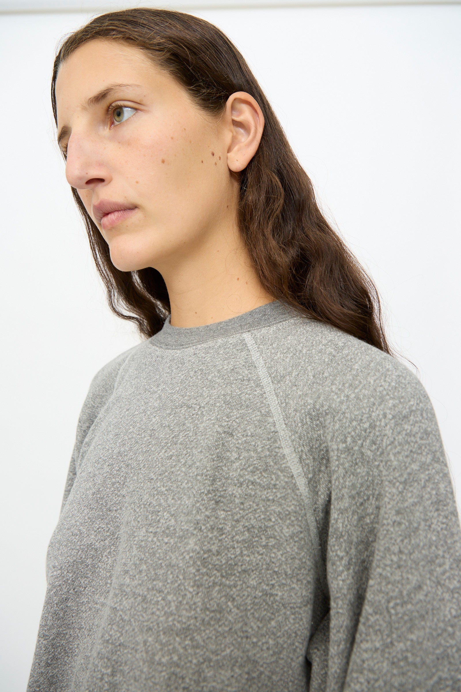 Model with long brown hair wears the B Sides Crewneck Sweatshirt in Heather Grey, looking slightly to the side against a plain white background.