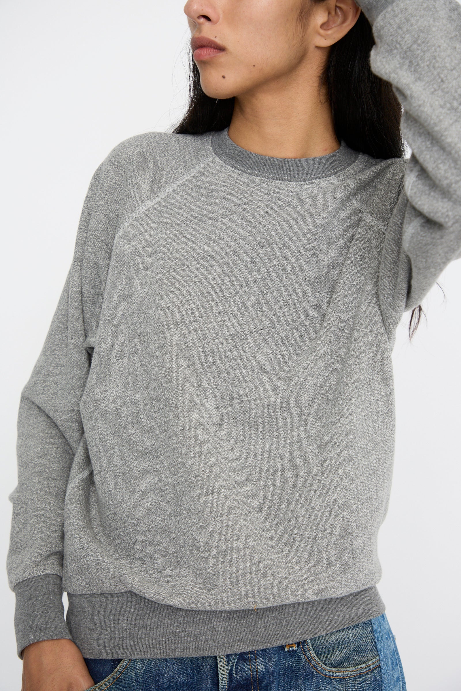 A person wearing a Crewneck Sweatshirt in Heather Grey by B Sides, styled with a shrunken collegiate fit, pairs it with blue jeans as they look to the side against a white background. 