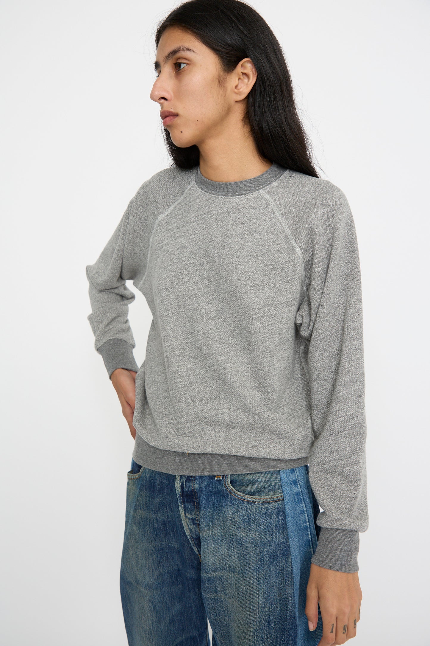 A person, in a Crewneck Sweatshirt in Heather Grey by B Sides with a shrunken collegiate fit and jeans, stands against a plain white background with a hand on their hip.