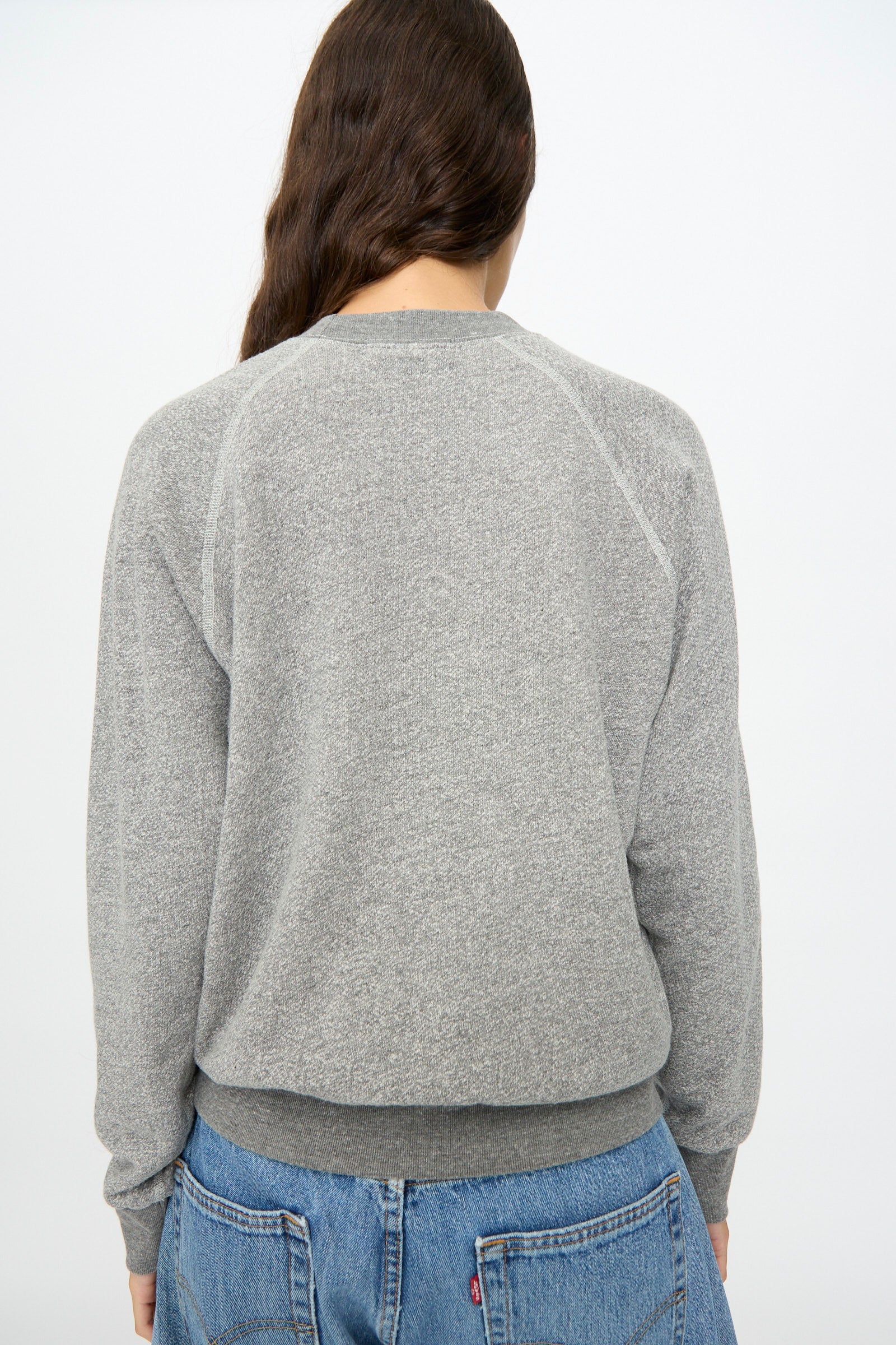 Individual with long brown hair shown from the back, wearing B Sides’ Crewneck Sweatshirt in Heather Grey and blue jeans against a plain light background.