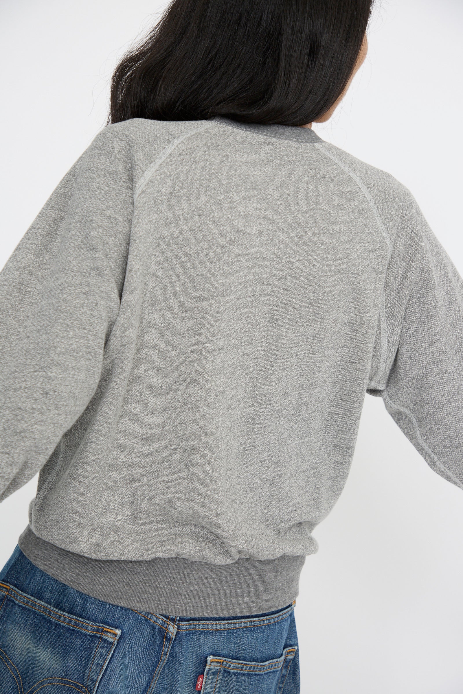 A person with long hair is wearing the B Sides Crewneck Sweatshirt in Heather Grey and blue jeans, facing away from the camera.