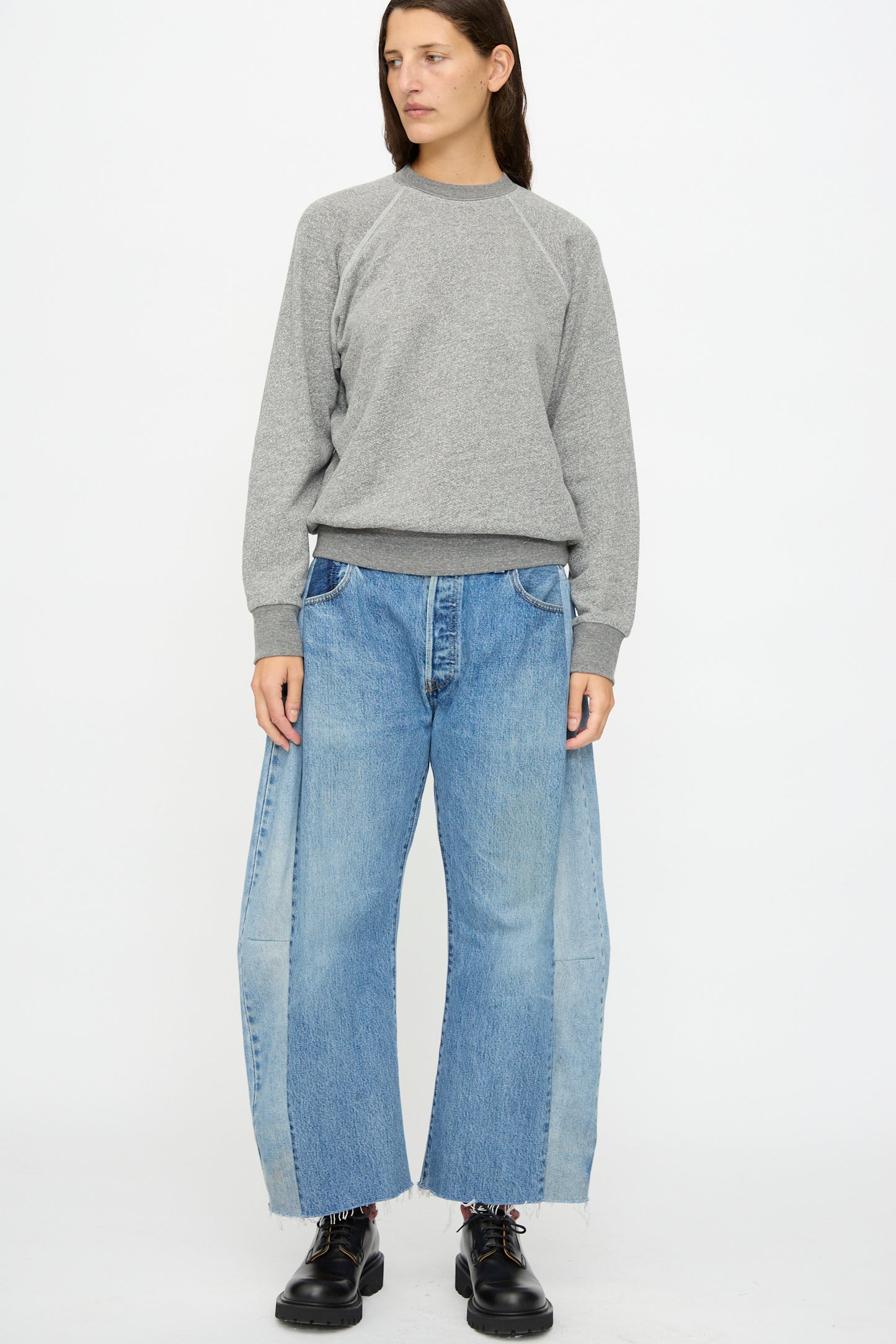 A person stands against a plain background wearing the B Sides Crewneck Sweatshirt in Heather Grey, wide-leg blue jeans with light panels, and black shoes.