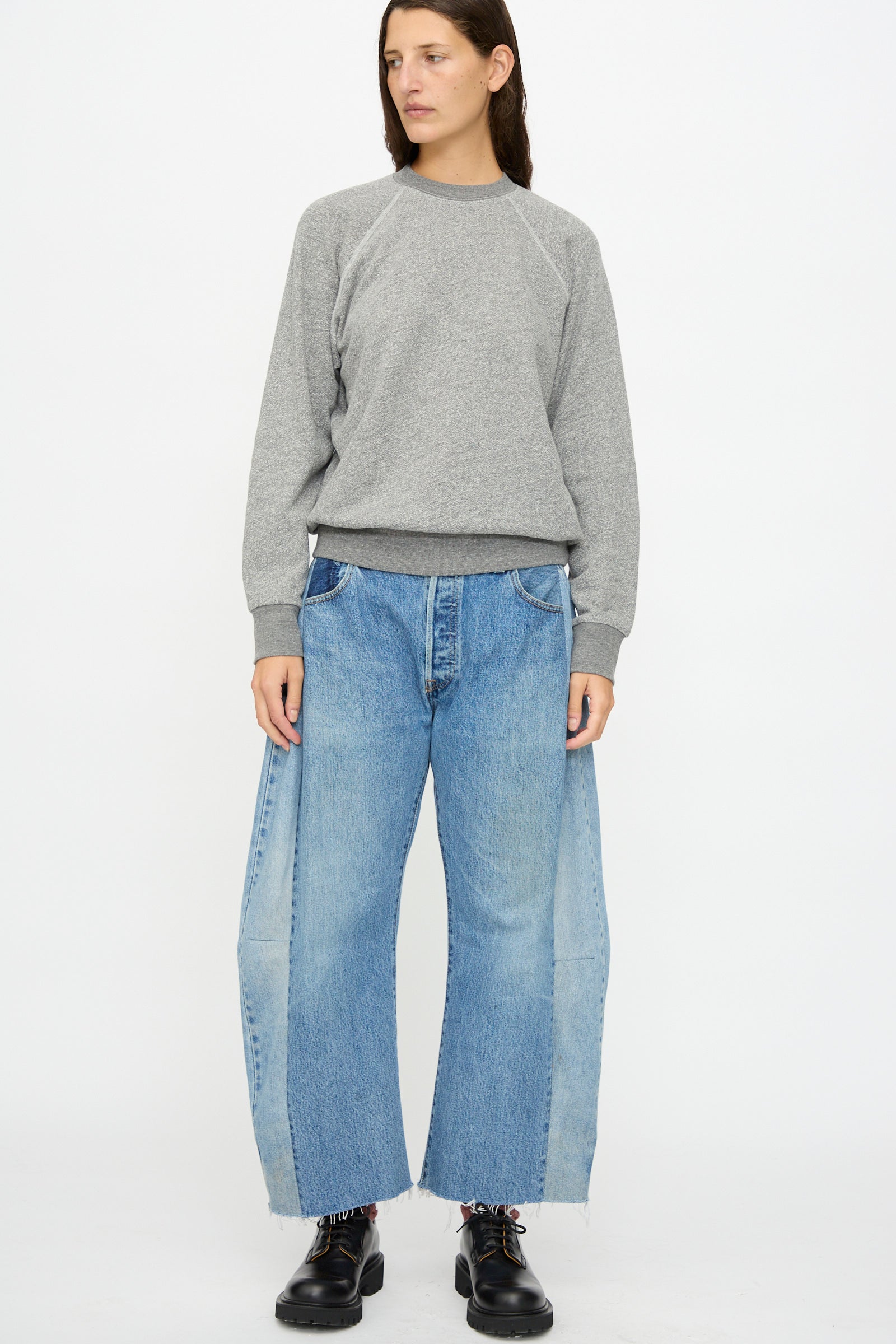 A person stands against a plain background wearing the B Sides Crewneck Sweatshirt in Heather Grey, wide-leg blue jeans with light panels, and black shoes.
