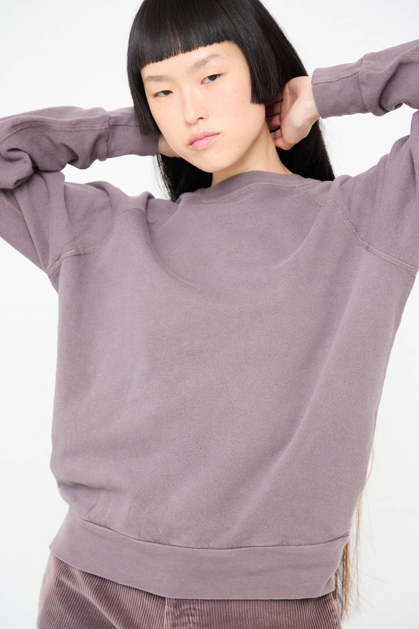 Person with long straight hair and blunt bangs wearing a loose-fitting B Sides Crewneck Sweatshirt in Tess Botanical stands with hands behind their head against a plain white background.