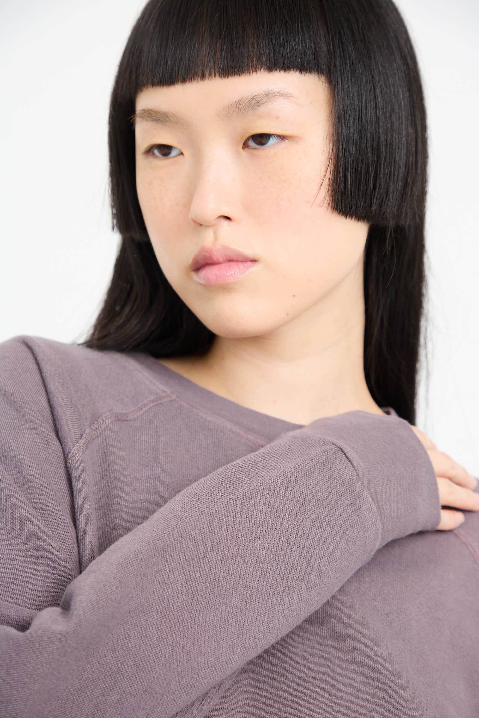 A person with long, straight, black hair and blunt bangs, wearing a mauve-colored Crewneck Sweatshirt in Tess Botanical by B Sides, looks off to the side.