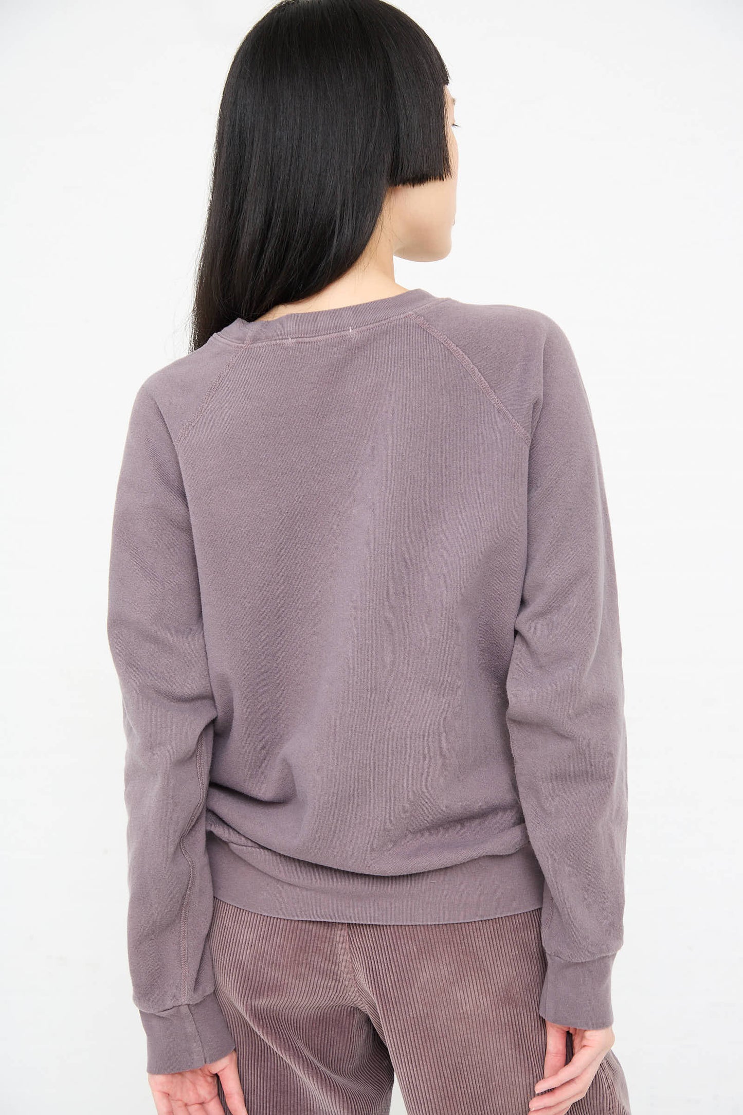 A person with long dark hair is standing with their back to the camera, wearing a Crewneck Sweatshirt in Tess Botanical from B Sides and light brown pants.