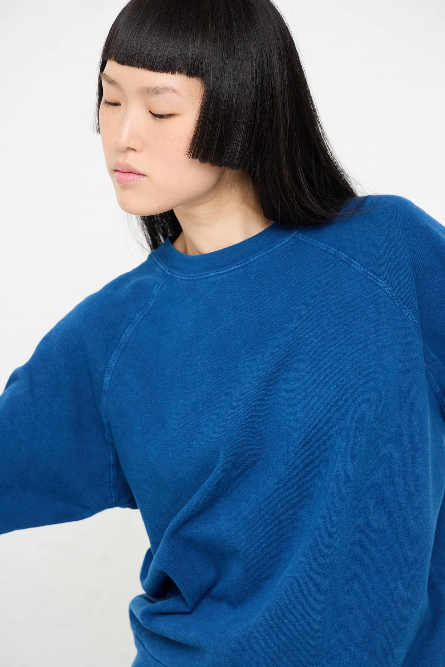 A person wearing a B Sides Crewneck Sweatshirt in Van Blue Botanical is looking down and to the side, with straight black hair and bangs, standing against a plain white background.