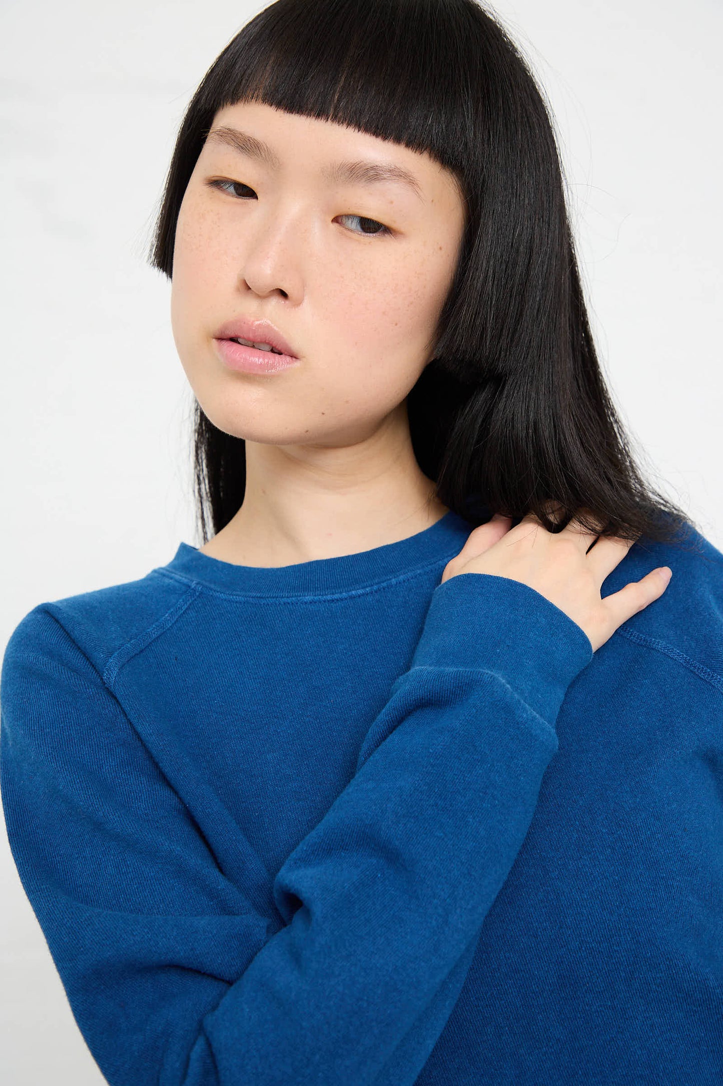 A person with long black hair wearing a B Sides Crewneck Sweatshirt in Van Blue Botanical touches their hair with one hand while looking slightly to the side.