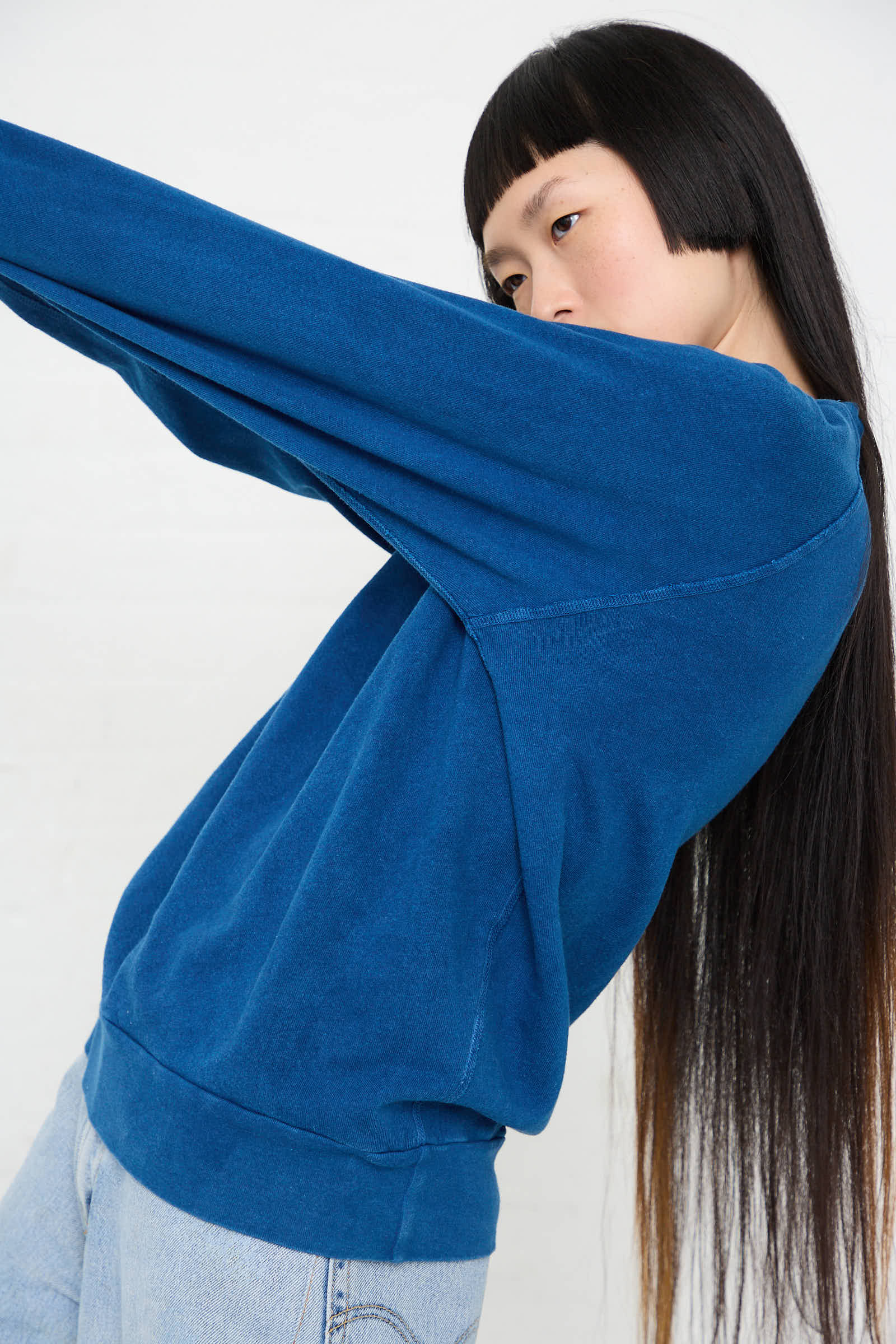 A person with long hair, wearing a B Sides Crewneck Sweatshirt in Van Blue Botanical and light blue jeans, is stretching their arms forward while slightly bending backward against a plain white background.