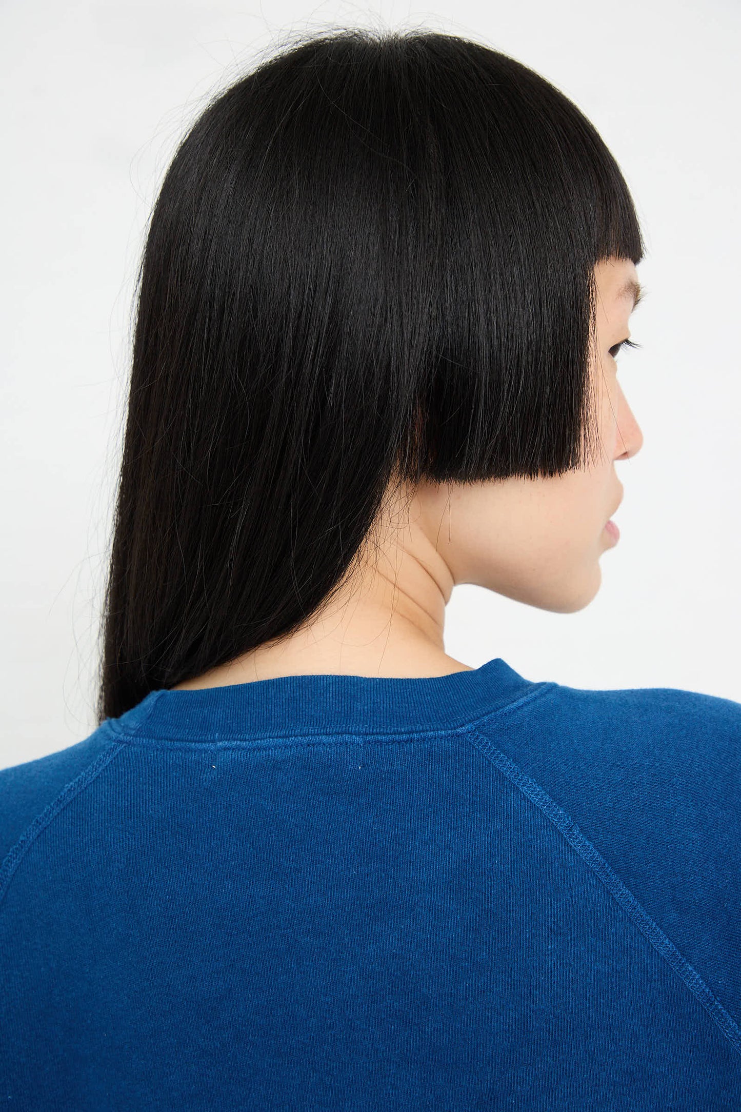 A person with straight black hair, a blunt bob haircut, and bangs is shown from the shoulders up, facing sideways, wearing the Crewneck Sweatshirt in Van Blue Botanical by B Sides.