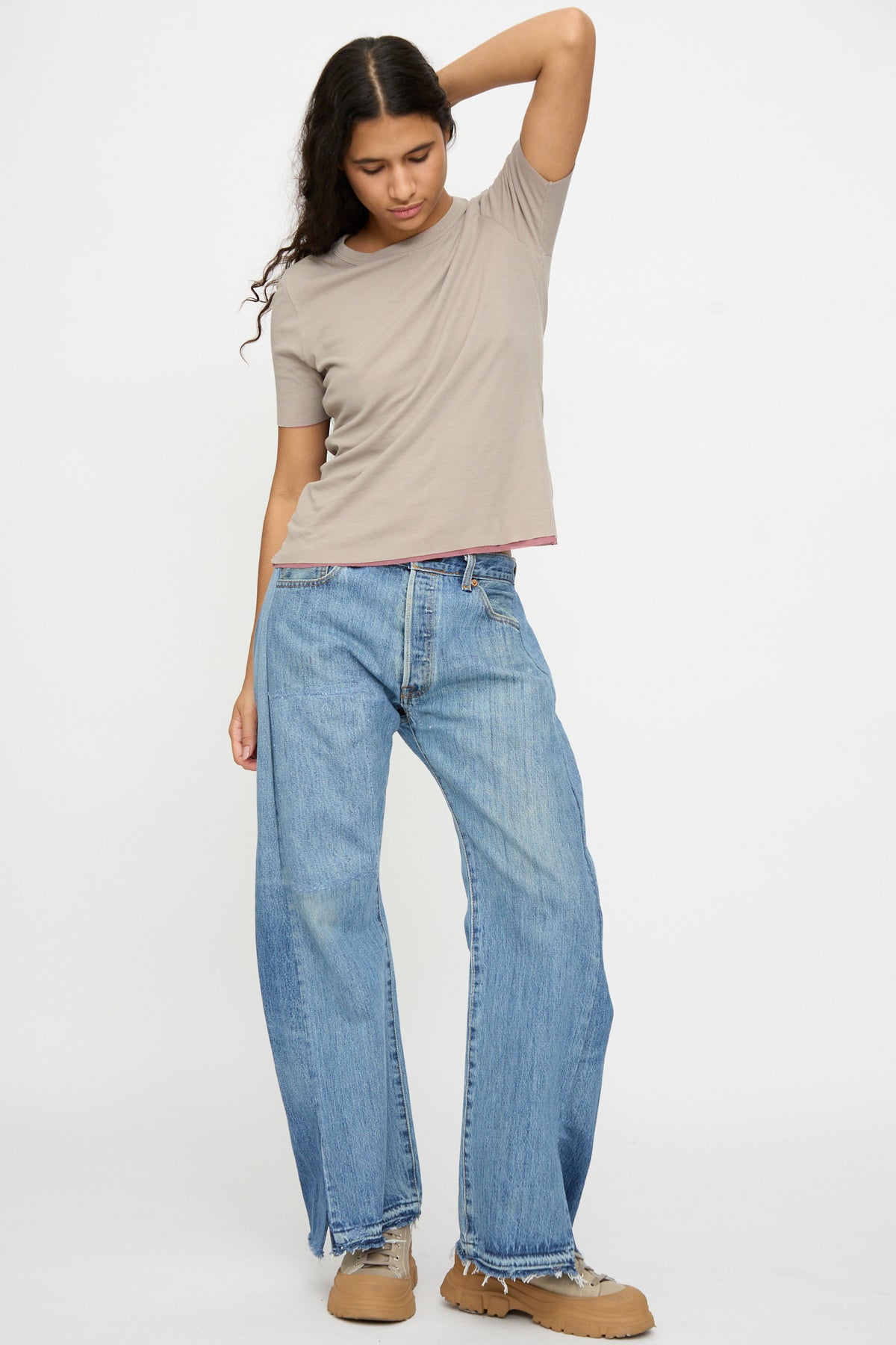 Person against a plain background, wearing the B Sides Double Tee in Pink and Dove Grey, relaxed fit blue jeans, tan shoes, with one arm raised and head slightly tilted.